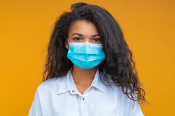 Close up studio portrait of young woman doctor wearing blue protecting surgical yellow mask on her face, isolated over yellow background