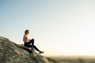 Woman hiker sitting on a steep big rock enjoying warm summer day. Young female climber resting during sports activity in nature. Active recreation in nature concept.