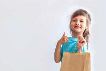 Smiling eight-year-old girl holds eco-friendly packaging in her hands, makes a hand gesture, light background, space for text
