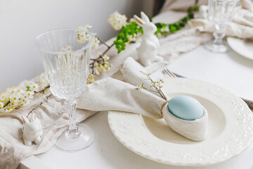 Stylish elegant Easter brunch table setting. Easter egg in bunny napkin on plate with cutlery, bunny, spring flowers on rustic white table. Modern Easter table arrangement