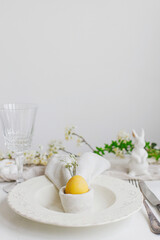 Easter egg in bunny napkin on plate with cutlery, bunny, spring flowers on rustic white table. Stylish elegant Easter brunch table setting. Modern table arrangement and eco decoration
