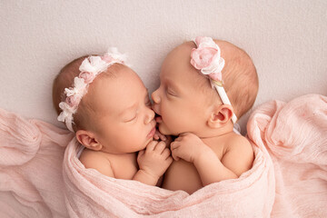 Tiny newborn twin girls. A newborn twin sleeps next to his sister. Newborn twin girls on the background of a pink blanket with pink bandages. The girls gently hug and kiss their sister in a cute pose. - obrazy, fototapety, plakaty