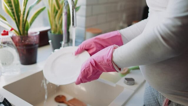 Pregnant woman wearing pink gloves washing dishes at home