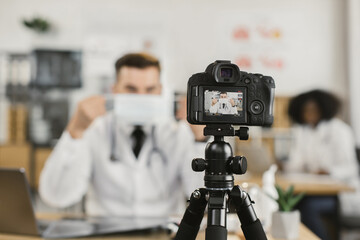 Competent caucasian doctor doing live streaming and explaining how to wear medical face mask. Medical specialist using modern laptop and digital camera for work.