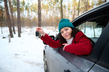 Beautiful Caucasian woman shows car keys through the driver's door window, smiling cutely while traveling in the winter snowy season. Safe driving and car travel concept