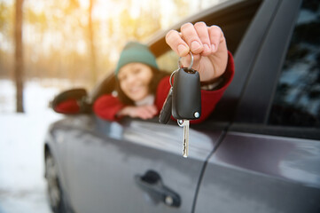 Focus on car key in the hand of a smiling female driver looking at camera against the background of a snowy nature with beautiful sunbeams falling into the forest on a beautiful sunny winter cold day
