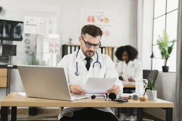 Focused caucasian doctor in eyeglasses and lab coat examining patient diagnosis during video appointment on modern laptop. African american nurse working on background.