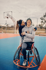 Happy athletic woman embraces her male friend with disability who is using wheelchair on basketball court.