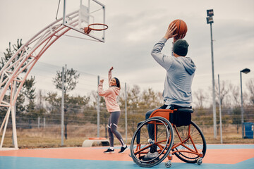 Wheelchair bound man shooting at the hoop while playing basketball with female friend outdoors.
