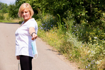 Fototapeta premium Beautiful blonde fitness woman in sports clothes standing sideways on the path in the forest stretched her hand forward holding the medical mask.