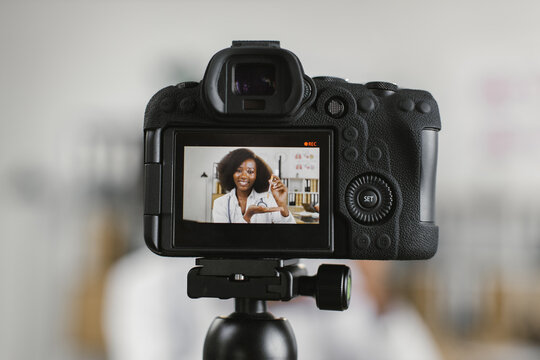 View through video camera on positive african american female doctor sitting at desk with sterile syringe in hands. Skillful medical worker talking about injection during vlog recording.