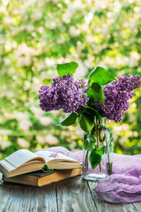 Romantic spring morning. A bouquet of purple lilacs and an open book on a wooden table overlooking the window. Still life concept, blurred background.