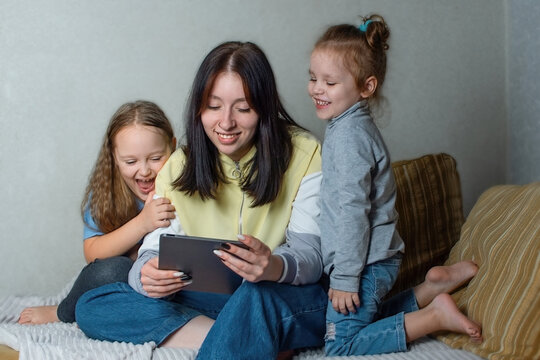 The Older Sister Is Talking And Watching Something Interesting On The Tablet, The Younger Two Sisters Are Interested In Watching What Is On The Screen Of The Gadget. Family Fun At Home