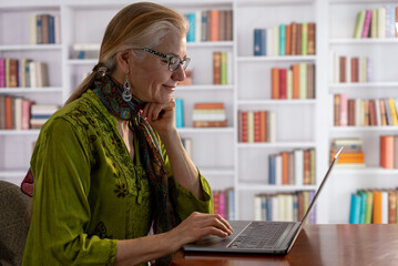 Profile of pretty, mature smiling woman working at a laptop computer in a home office library.