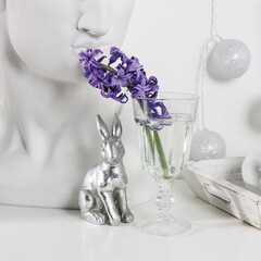 Ceramic figurines of Easter bunnies of different sizes on the table. Blue and white hyacinths in glass cups on a white background. Easter design