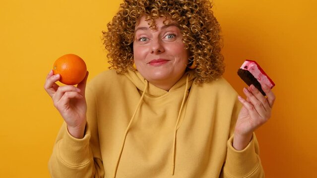 Portrait of pretty young fat woman looking at camera choosing between orange and cream cake isolated yellow color background. Healthy dieting and eating habits concept. Losing weight on diet