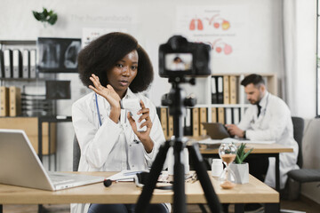 Fototapeta premium African female practitioner recording video blog about usage of modern infrared thermometer while her caucasian male assistant working on laptop behind. Concept of medicine and technology.