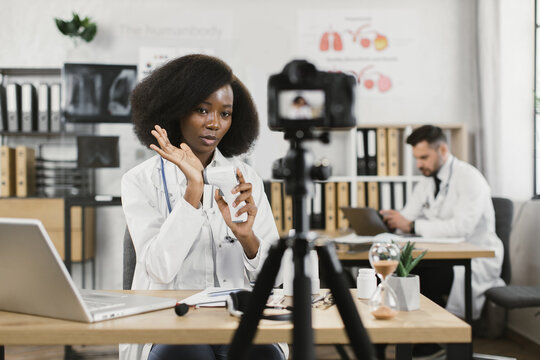 African female practitioner recording video blog about usage of modern infrared thermometer while her caucasian male assistant working on laptop behind. Concept of medicine and technology.