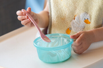 cute hands of a little hungry caucasian baby or toddler wearing a soft yellow napkin eating white Jogurt out of a blue plastic cup. Small kid learning to eat food by itself while growing up.