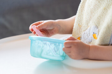 cute hands of a little hungry caucasian baby or toddler wearing a soft yellow napkin eating white Jogurt out of a blue plastic cup. Small kid learning to eat food by itself while growing up.