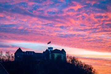 Castle of Ljubljana located on a hill surrounded by trees under a dramatic glowing sky with pink and orange illuminated clouds at sunset or sunrise on a beautiful day. Flags swaying in the wind.