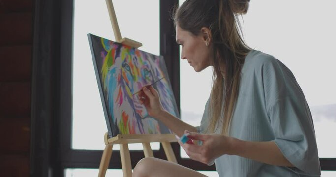 Hobby at home during self-isolation, woman artist is painting picture on canvas near window in wooden house. Slow motion