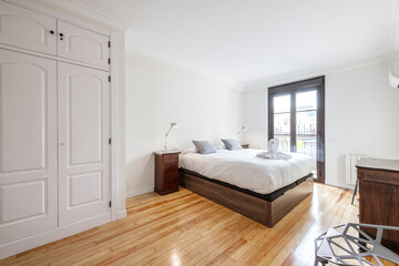 Fototapeta na wymiar bedroom with a dark wooden bed, white lamps on the bedside tables, a built-in wardrobe and a large window with a balcony in the background
