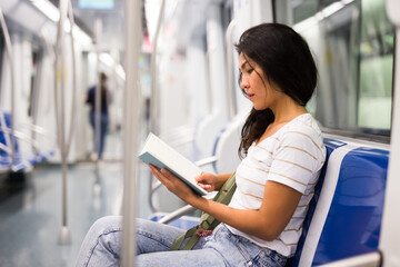 Asian woman sitting on bench in subway train and reading notes in her notebook.