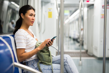 Asian woman with bag and sitting on bench in subway train and using smartphone.