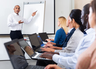 Portrait of hispanic business coach giving talk before participants of corporate training sitting with laptops in meeting room