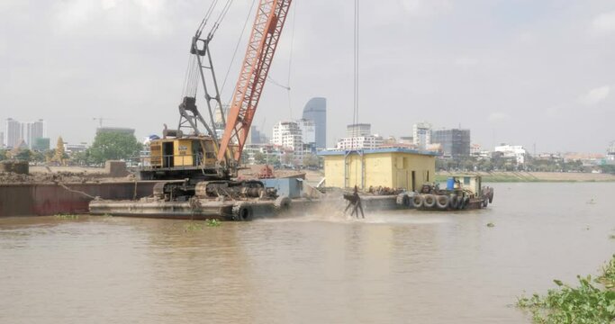 close up of a grab dredger in operation. Dredged sand is loaded into the barge. Phnom Penh capital as backdrop