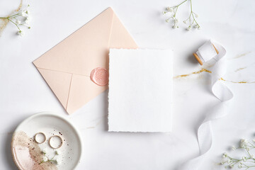 Elegant wedding stationery set. Flat lay wedding invitation card template, pastel pink envelope, silk ribbon, golden rings, flowers on marble desk.