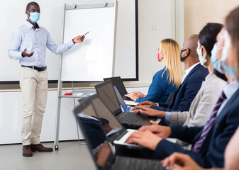 Young african american businessman in protective mask to prevent viral infection presenting business project to partners sitting with laptops in conference room