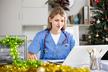 Young positive female doctor sitting in office at workplace and working at laptop during Christmas time