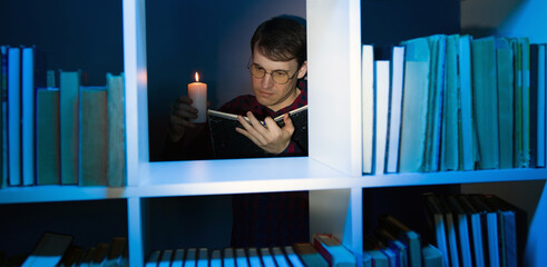Man in glasses with candle reading book, standing behind bookshelf. Male holds burning candle while reading book in wooden bookcase with various literature in dark room.