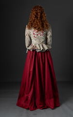 A mannequin with long red, curly hair and wearing a renaissance-style embroidered bodice and a red skirt and shown from behind
