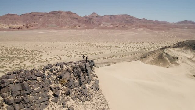Two guys atop rocky outcrop next to high dune in Namibian wilderness; aerial