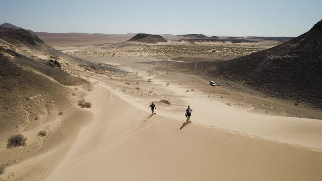 Two travelers walk down big sand dune, unforgiving desolate terrain; drone