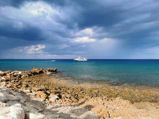 Fototapeta na wymiar The stone coast of the Mediterranean Sea against the backdrop of the sea with a walking white ship and a dramatic sky.