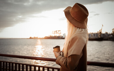 Stylish blonde woman in felt hat and coat drinks coffee on the go at sunrise at sea