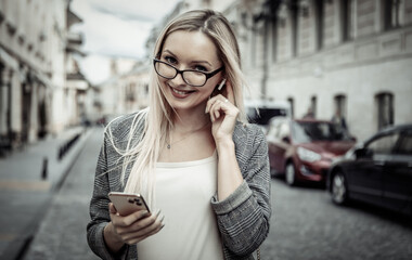 Portrait of confident business woman in urban street. Blond girl in business suit using smartphone and listening to music with wireless headphones