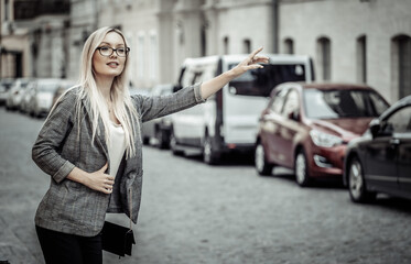 Naklejka na ściany i meble Beautiful Woman Catching Taxi Car On Street. Portrait Blonde Business Woman On Way To Work Stopping Taxi Car Outdoors.