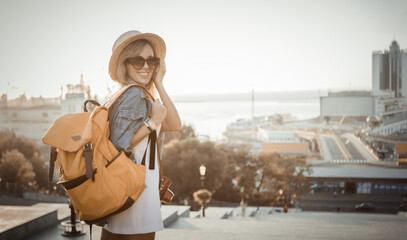 Portrait of young smiling woman tourist with a big backpack in the city at sunrise. Travel concept - obrazy, fototapety, plakaty