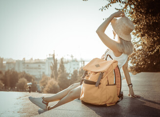 Cute woman tourist sits in the city with large travel backpack in the rays of the sun at sunrise - obrazy, fototapety, plakaty