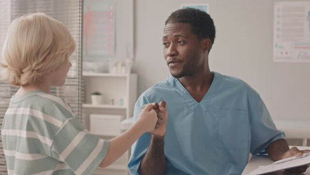 Medium slowmo shot of little Caucasian boy coming to doctors office. African-American young male pediatrician greeting him with fist bump