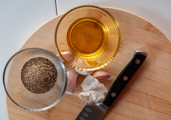 A glass with olive oil and a glass with black pepper with garlic and a knife on a chopping wooden board, as part of ingredients for cooking. top view or flat lay image