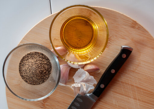 A glass with olive oil and a glass with black pepper with garlic and a knife on a chopping wooden board, as part of ingredients for cooking. top view or flat lay image