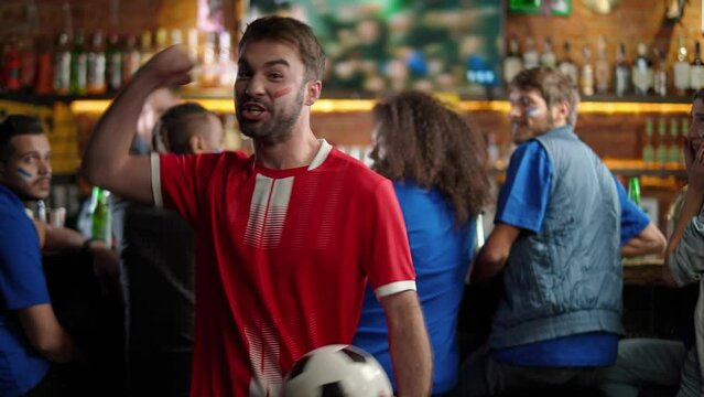 Football fans of different races drink beer and watching a football match at the bar, fan of the opposite team enjoy scoring a goal during a football match and rejoices at the victory, funny moment.