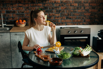 Young woman preparing vegetarian greens salad at home and tasting yellow pepper
