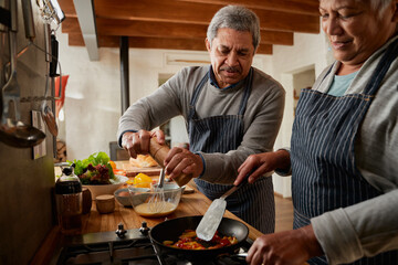 Elderly multi-ethnic couple cooking breakfast together in modern kitchen. Husband grinding spices...
